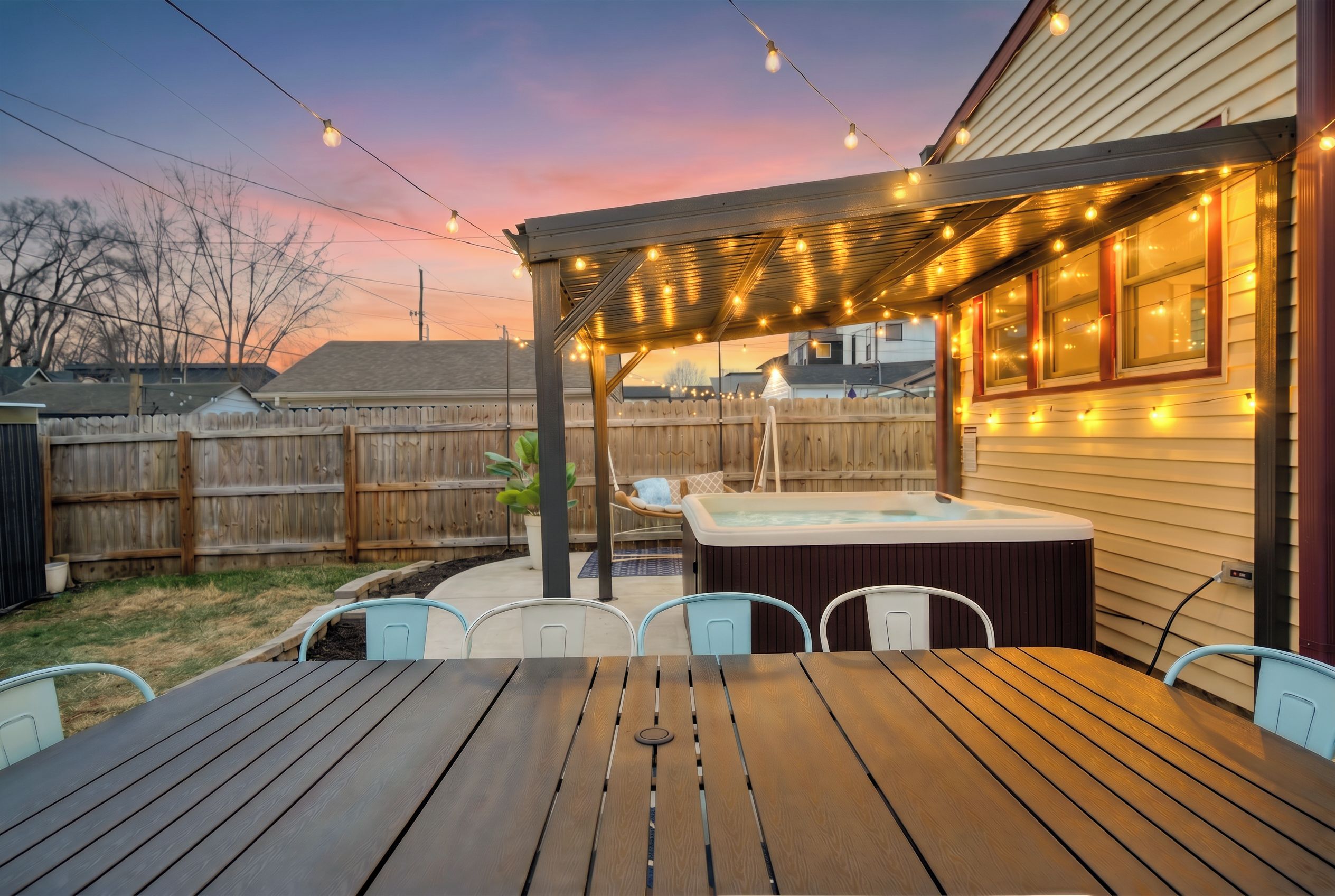 A backyard patio with a wooden table and chairs, a hot tub, and string lights.