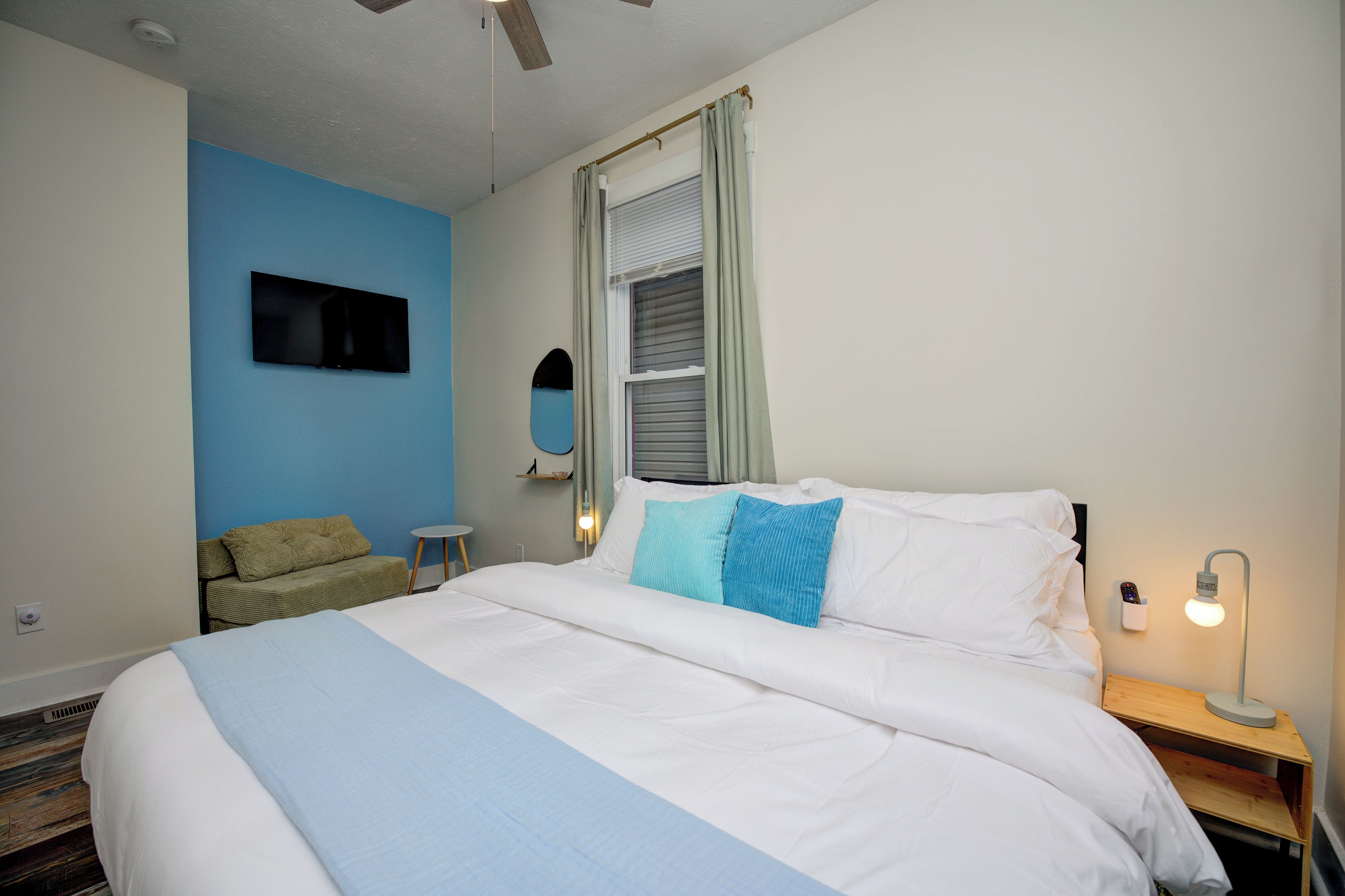 Een hotelkamer met een bed, een aan de muur bevestigde televisie, een klein tafeltje, een nachtkastje met een lamp en een raam met gordijnen.