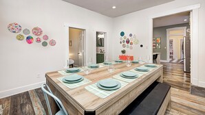 Zona pranzo con un lungo tavolo in legno apparecchiato per sei persone, circondato da sedie. Si notano piatti decorativi appesi alle pareti e si intravede una cucina attraverso una porta aperta.