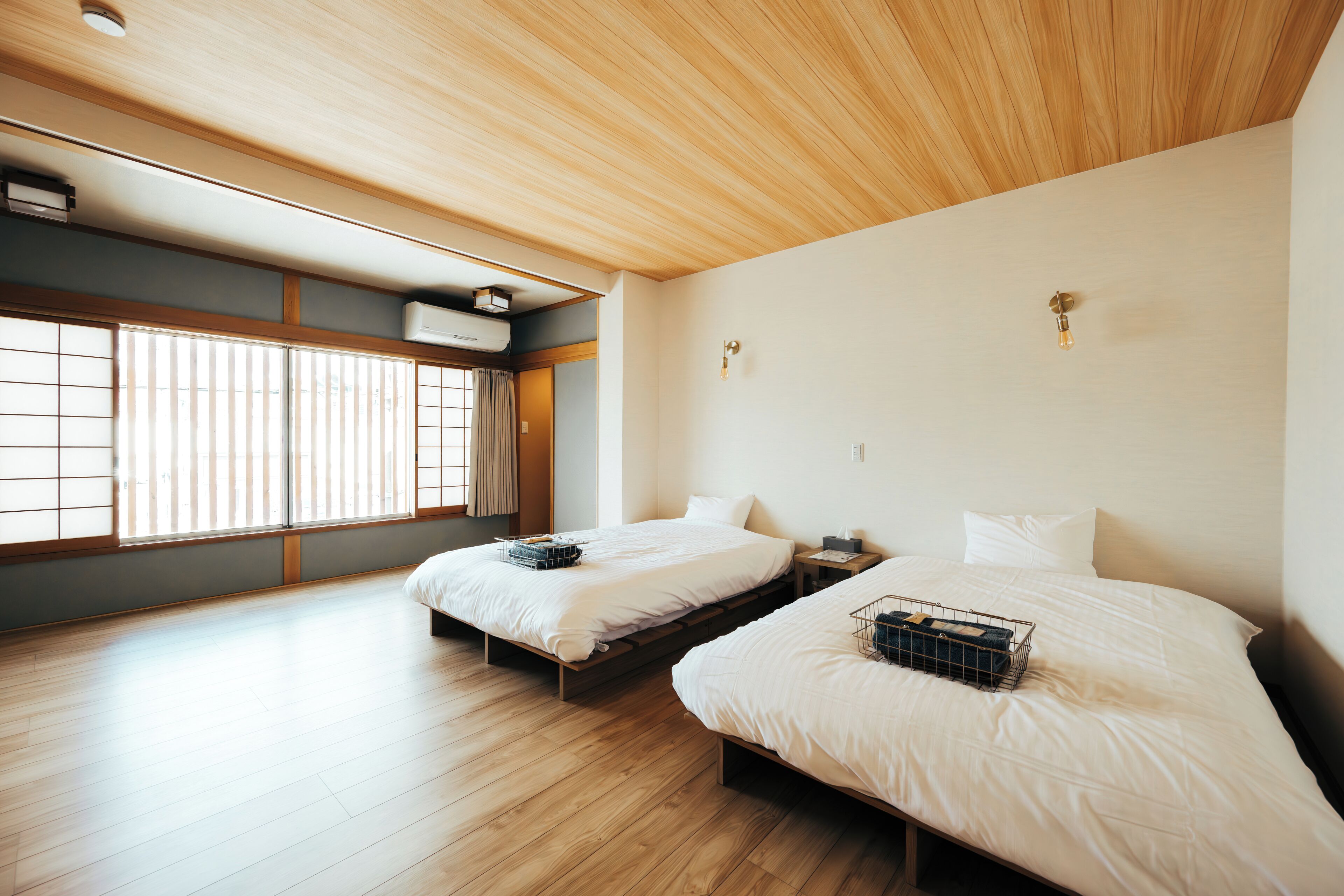 Ein Zimmer mit zwei Betten, Holzboden, einer Decke mit Holzzaune und einem Fenster mit Jalousien.