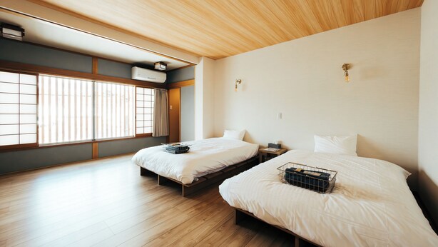 Una habitación con dos camas, suelo de madera, techo con paneles de madera y una ventana con persianas.