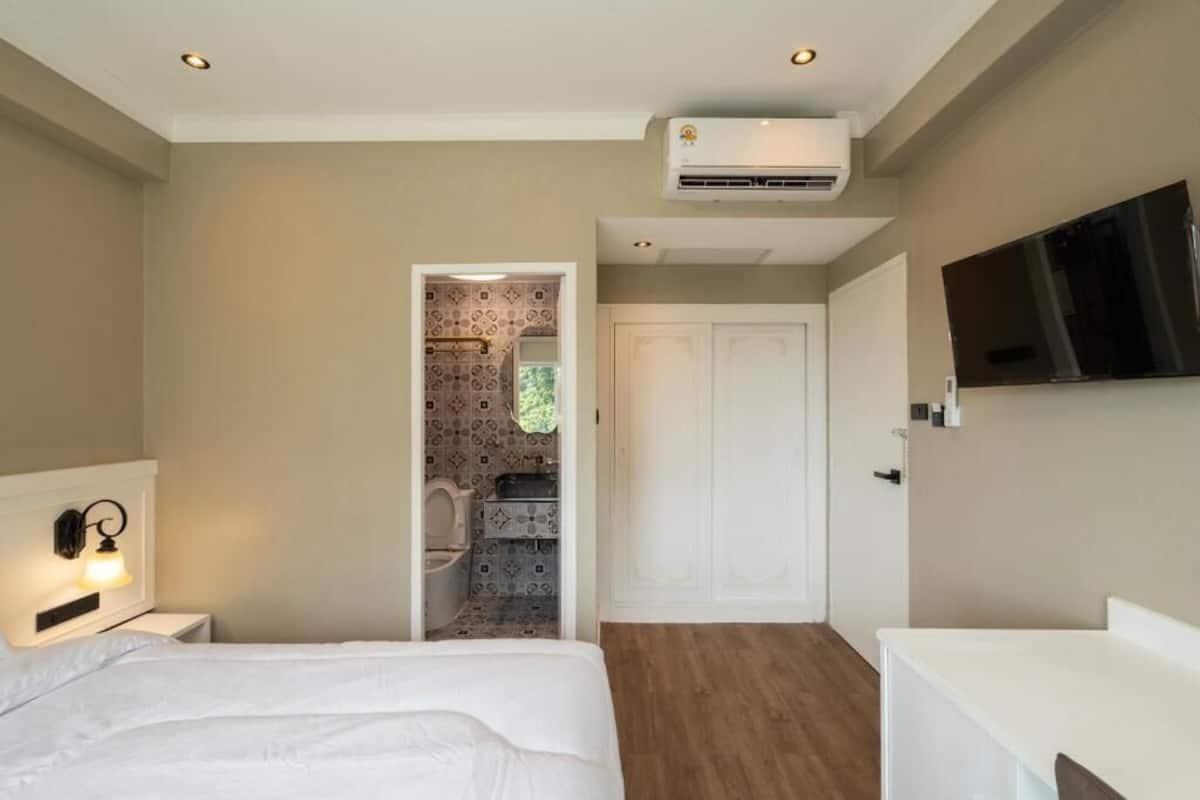 A bedroom with a bed, a white bedside table, a wall-mounted TV, and an air conditioning unit.