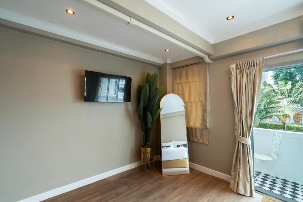 A room with a flat-screen TV mounted on the wall, a large mirror, a potted plant, a window with beige curtains, and a balcony with a checkered floor.