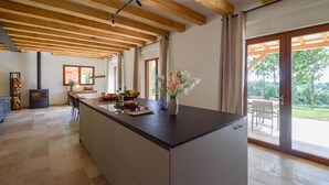 Eine geräumige Küche mit einer großen Kochinsel, Holzbalken an der Decke und Blick ins Freie durch große Schiebeglastüren.
