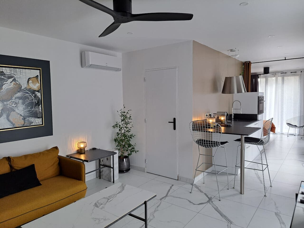 Una sala moderna con un sofá, una zona de comedor con mesa y sillas, un ventilador de techo y una puerta que da a otra habitación.