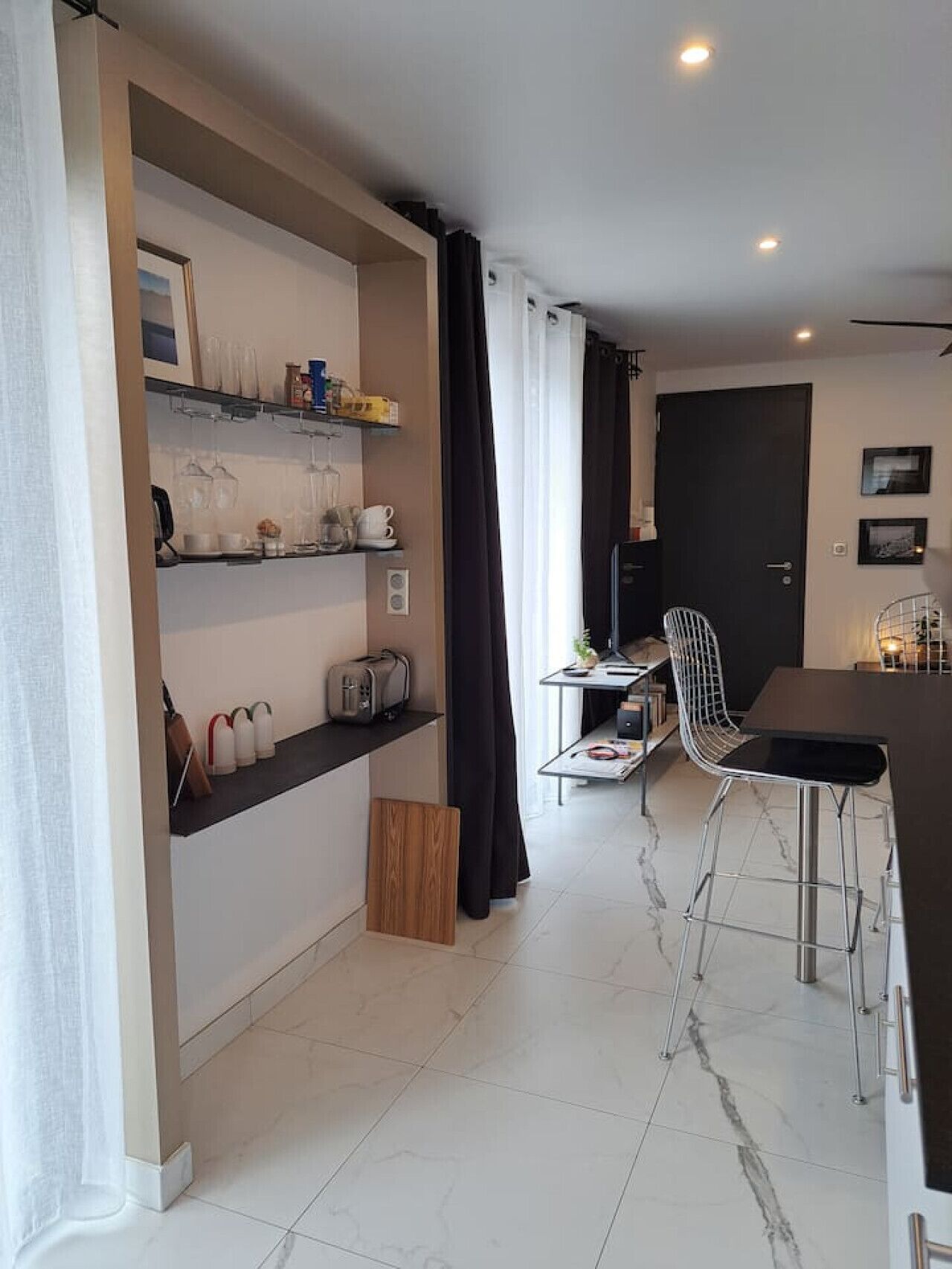 A compact kitchen with a dining area, featuring a built-in shelf with kitchenware, a black curtain, and a modern chair.