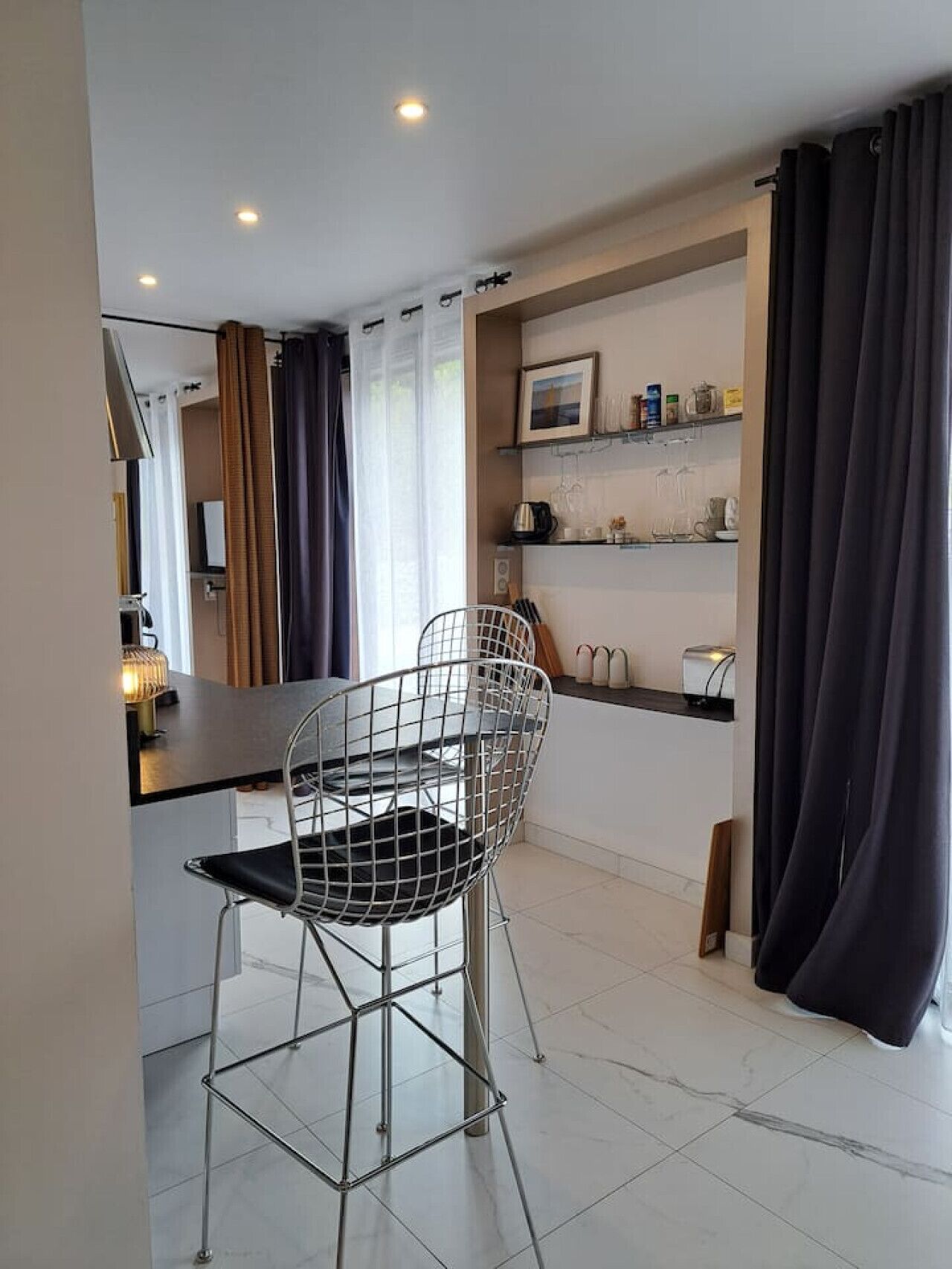 A modern kitchen with a dining area, featuring a metal wire chair, a small table, and a shelf with kitchen items.
