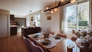 A dining area with a wooden table set for four, a kitchen in the background, and a large window with a view of greenery. - Holiday home for 6 people in the heart of Perigord (Saint-Médard-d’Excideuil)