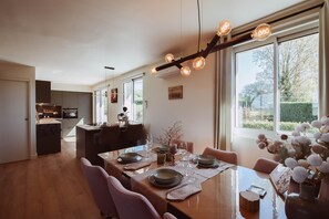 Un comedor con una mesa de madera dispuesta para cuatro, una cocina al fondo y un gran ventanal con vistas a la vegetación.