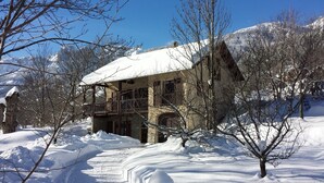 Un bâtiment en pierre dont le toit est recouvert de neige, entouré d’arbres et de montagnes également enneigés, qui se dressent à l’arrière-plan.
