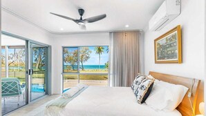 A bedroom with a bed, a ceiling fan, a large window with a beach view, and an air conditioning unit. - Luxury beachfront escape: Ocean views and poolside living! (Grasstree Beach)