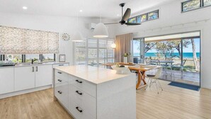 A modern kitchen with white cabinetry, a central island, pendant lights, and a view of the beach through large windows. - Luxury beachfront escape: Ocean views and poolside living! (Grasstree Beach)