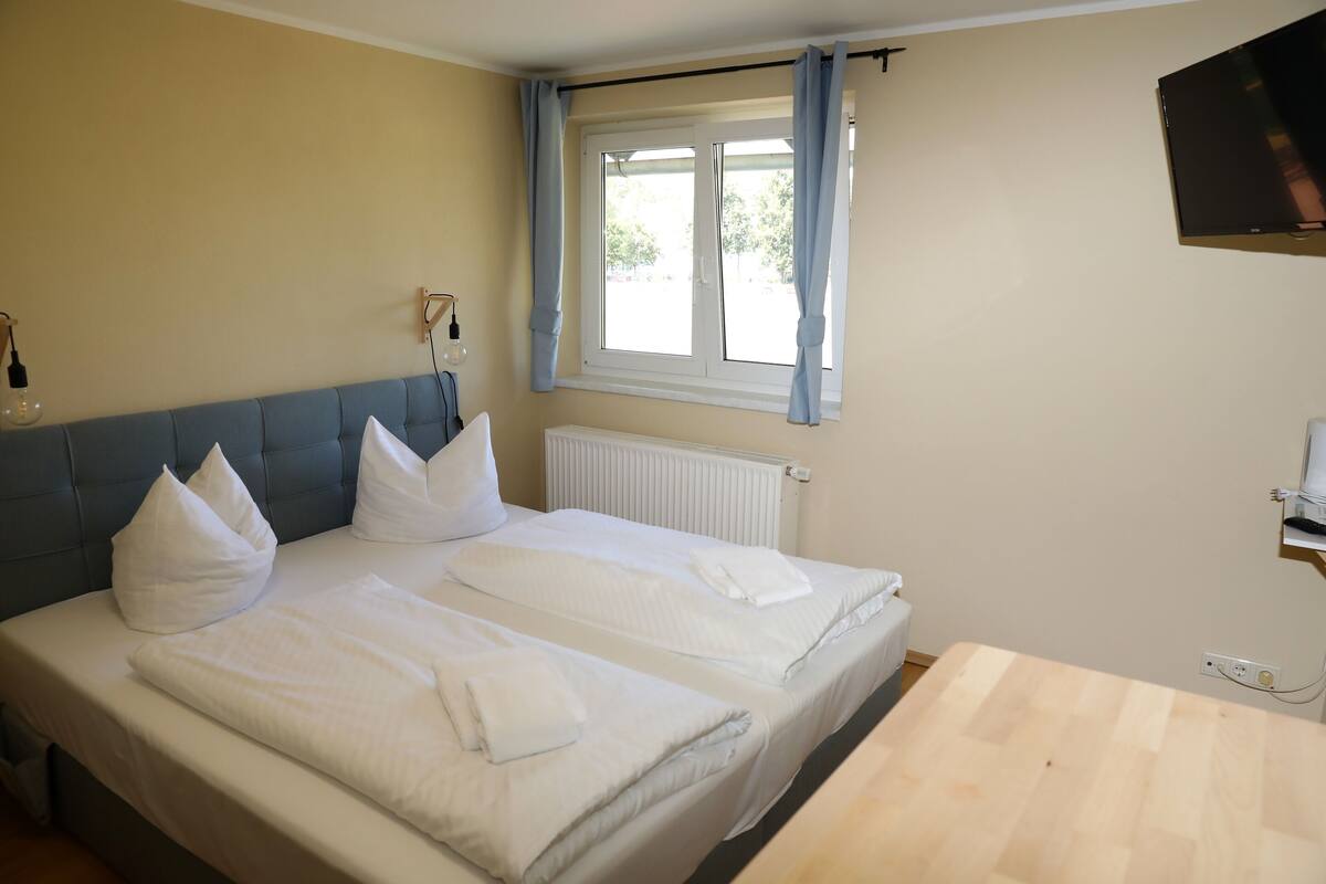A double bed with white linens, a wooden headboard, a window with curtains, a radiator, and a TV mounted on the wall.