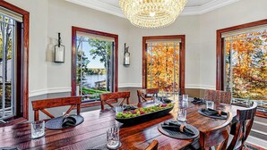 Ein Esszimmer mit Holztisch, Stühlen, einer Chandelüster und großen Fenstern mit Blick auf Herbstlaub.