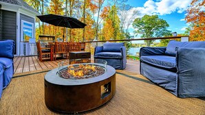 Eine Terrasse mit Gartenmöbeln, einem Feuerkorb und Blick auf Bäume und einen See.