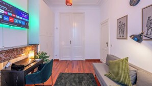 A modern living room with a flat-screen TV displaying streaming services, a desk with a lamp, a sofa with cushions, and a door in the background. - Room-euro Edens Brussels - Cozy Family S (Brussels)