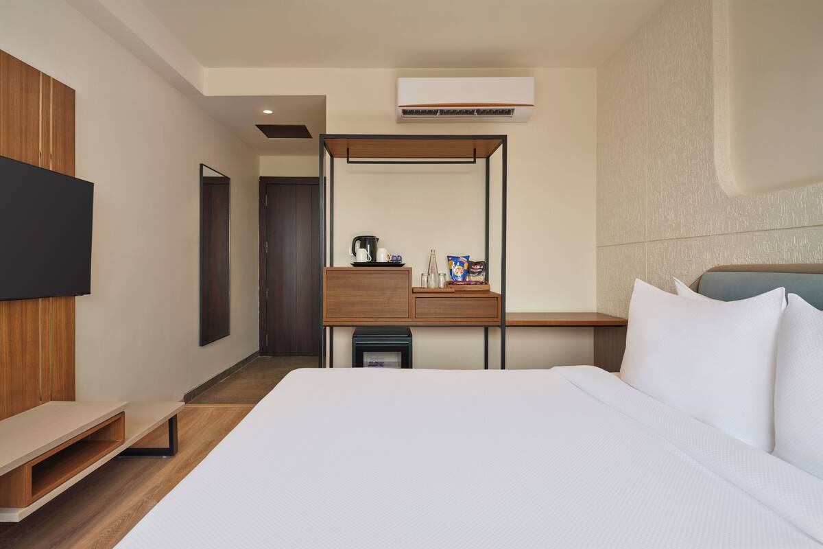 A modern hotel room with a large bed, a flat-screen TV mounted on the wall, a wooden desk with a coffee maker, and an air conditioning unit.
