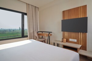 A hotel room with a large bed, a desk, a chair, a TV mounted on the wall, and a window with a view of greenery.