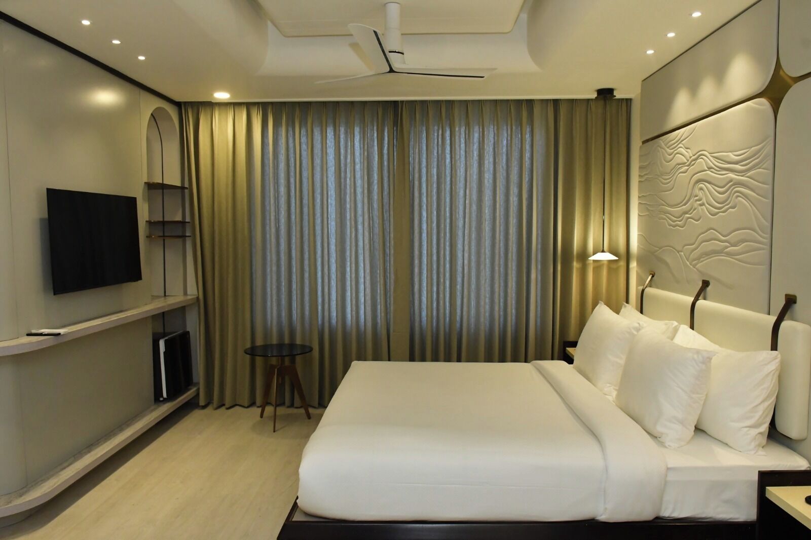 A modern hotel room with a large bed, a flat-screen TV mounted on the wall, a small round table, and a ceiling fan.