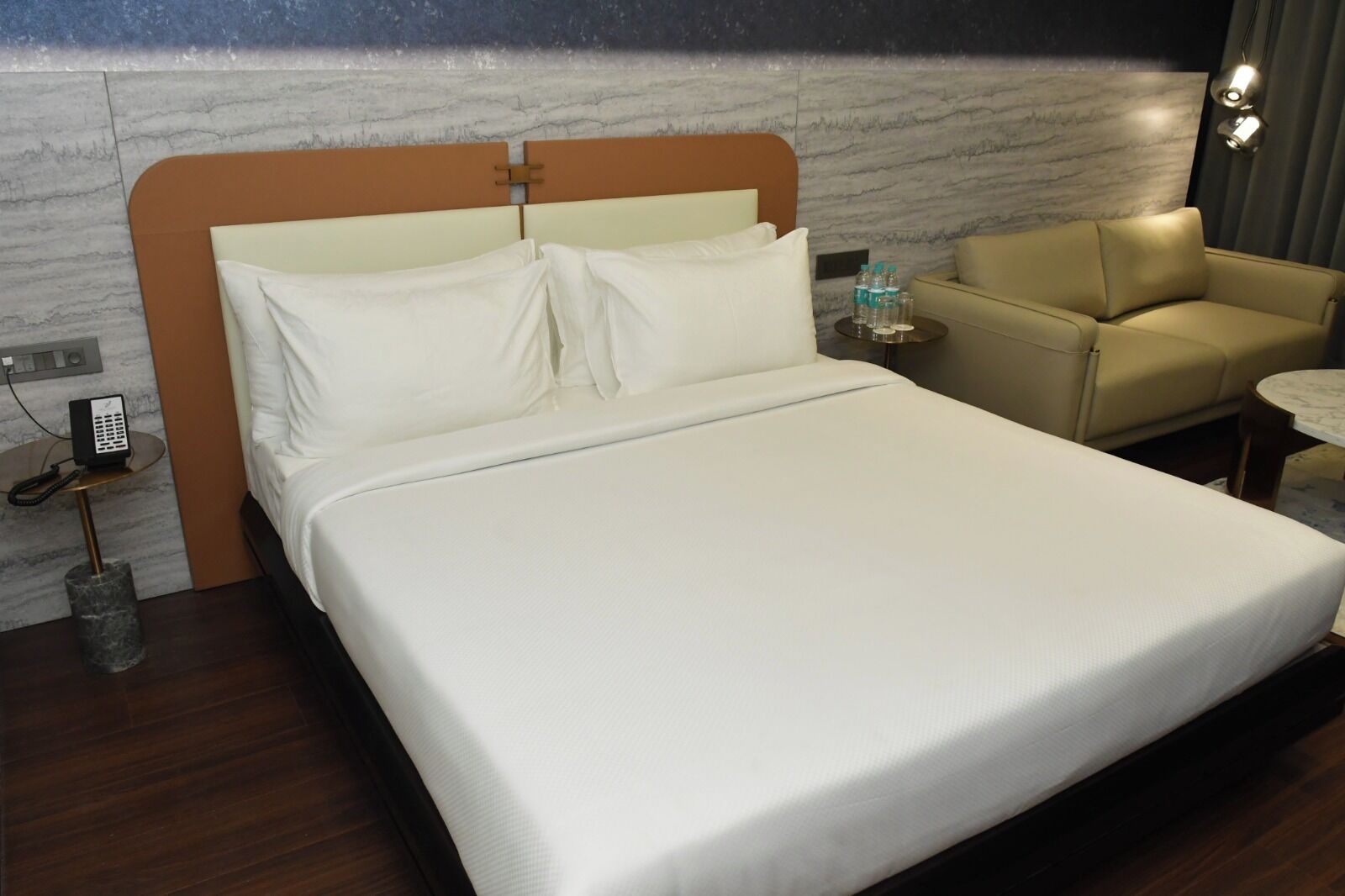 A hotel room with a large bed, a bedside table with a phone, a chair, and a small table with water bottles.