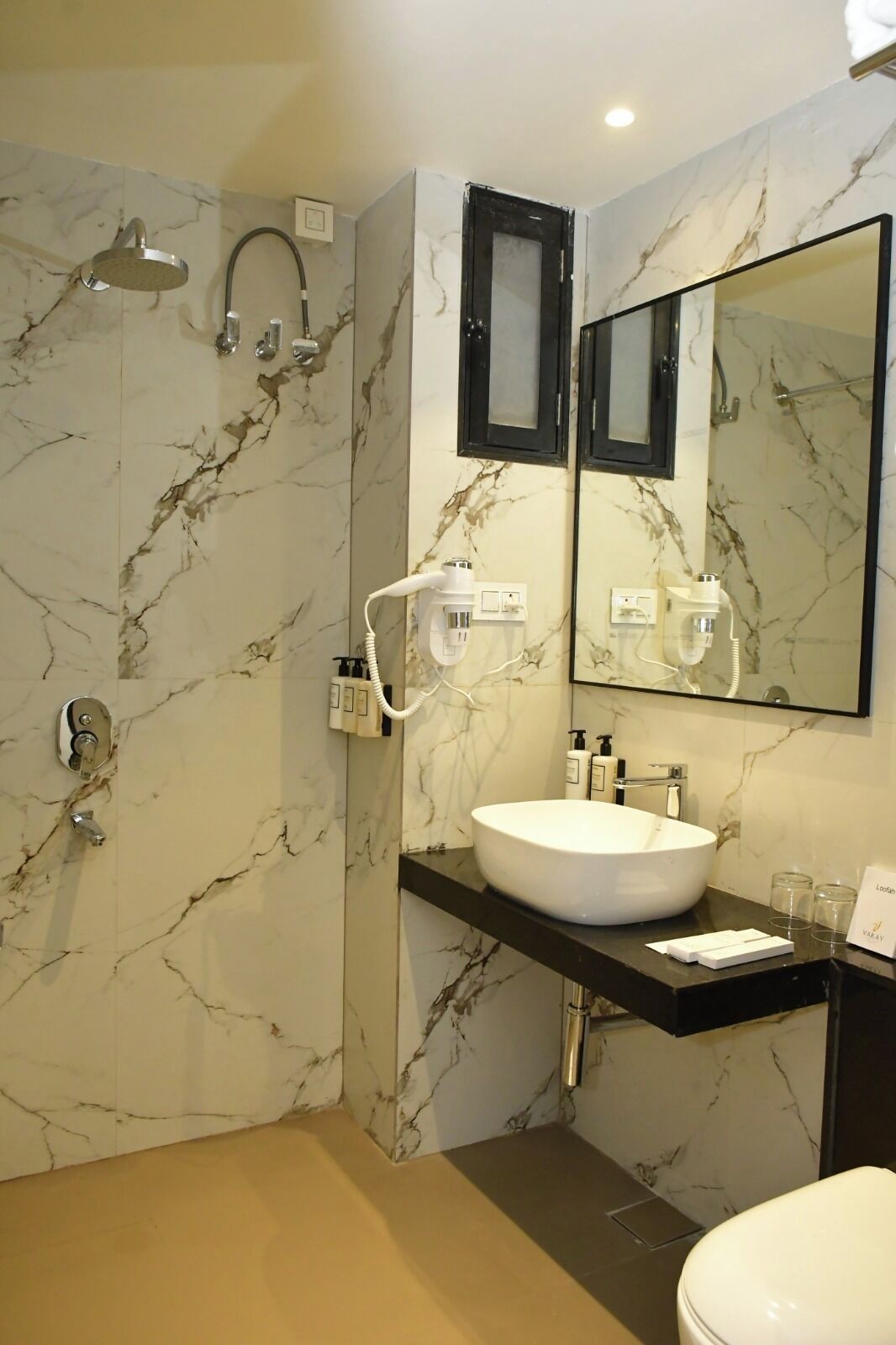 Un baño moderno con pared de mármol, un espejo grande, un lavamanos blanco, ducha y retrete.