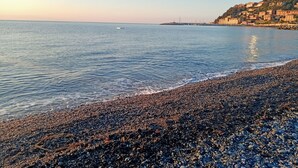 Pikkukivinen ranta, jossa on tyyni meri ja taustalla näkyy rakennuksia.