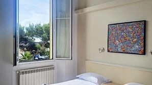 Hotellihuone, jossa on kaksi sänkyä, suuri ikkuna, josta avautuu näkymä vehreään maisemaan, patteri ja värikäs abstrakti maalaus seinällä.