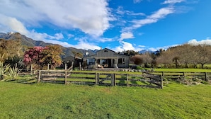Une maison moderne dotée d’une grande baie vitrée, entourée d’un jardin clos, avec des montagnes en arrière-plan.