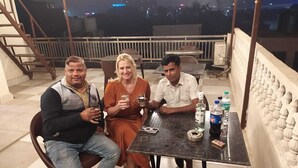 Tre persone sedute a un tavolo su un balcone con ringhiera, scale e vista sulla città sullo sfondo.