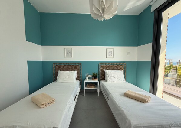 A hotel room with two beds, a small bedside table with a lamp, and a view of the outdoors. - Villa Flora's 3-BR Retreat & Private Pool in Protaras (Paralimni)
