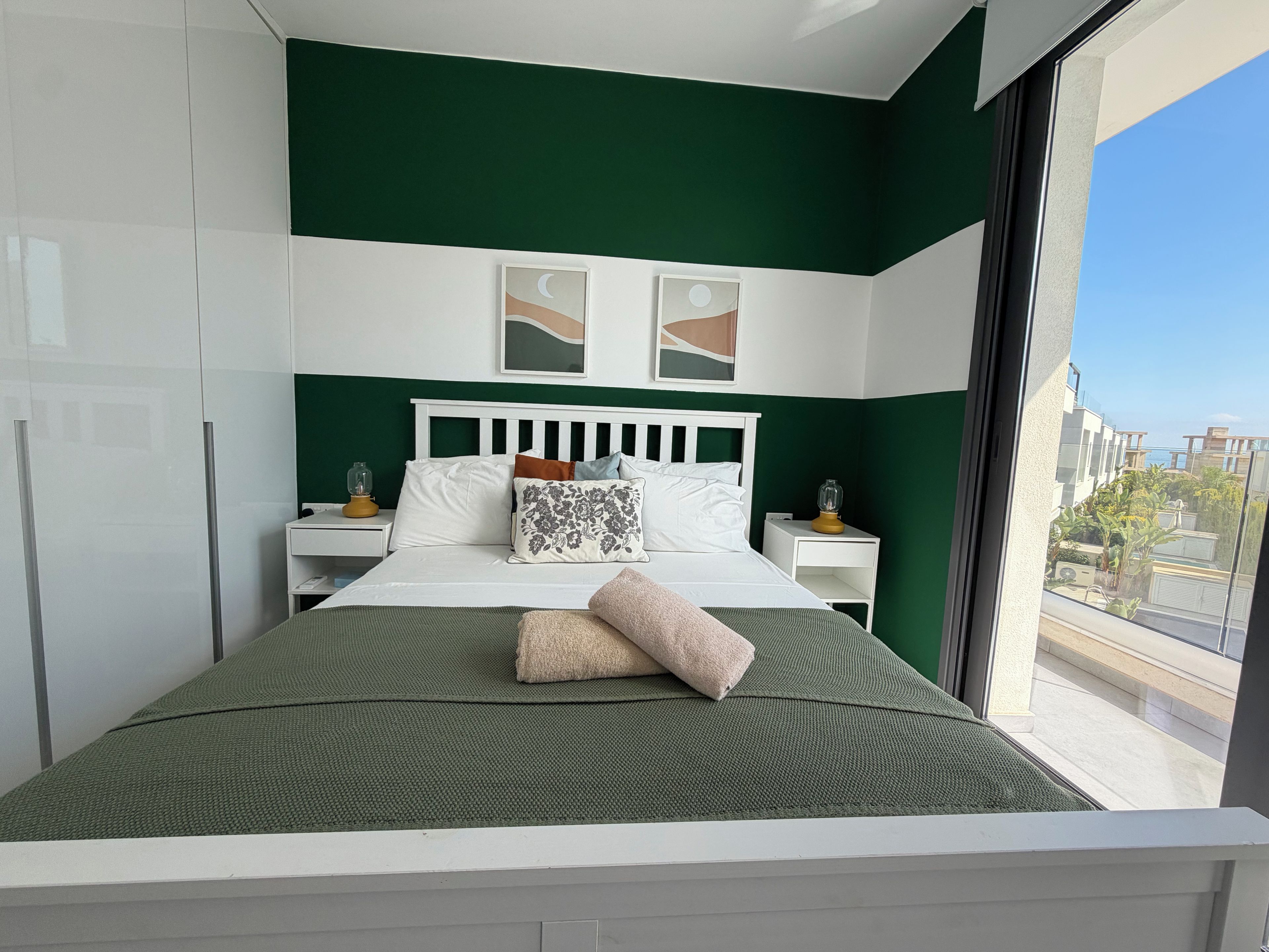A modern bedroom with a green and white color scheme, a bed with multiple pillows, a bedside table with a lamp, and a window with a view of buildings.