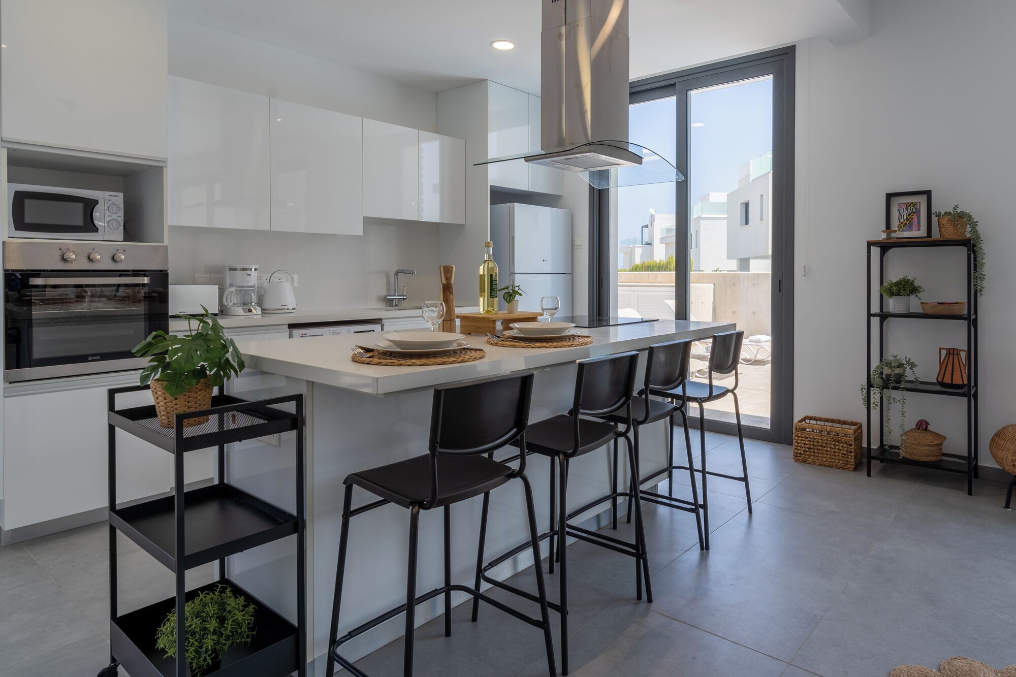 Moderni keittiö, jossa on valkoiset kaapit, keskellä sijaitseva saareke baarituoleineen, sisäänrakennettu uuni ja suuri ikkuna, josta avautuu näkymä ulos.