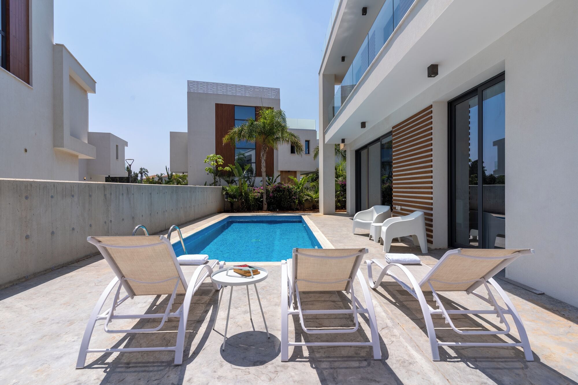 Uma área moderna de piscina ao ar livre com espreguiçadeiras e uma mesa, cercada por edifícios com grandes janelas e uma parede de concreto.