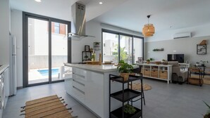 Une cuisine moderne dotée d’un grand îlot central, d’un coin repas avec un canapé et un téléviseur, et d’une piscine visible à travers les portes vitrées.