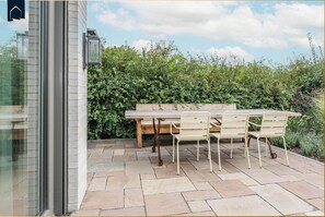 Eine Terrasse im Freien mit einem Holztisch und -stühlen, einer Bank und einem Hecken im Hintergrund.