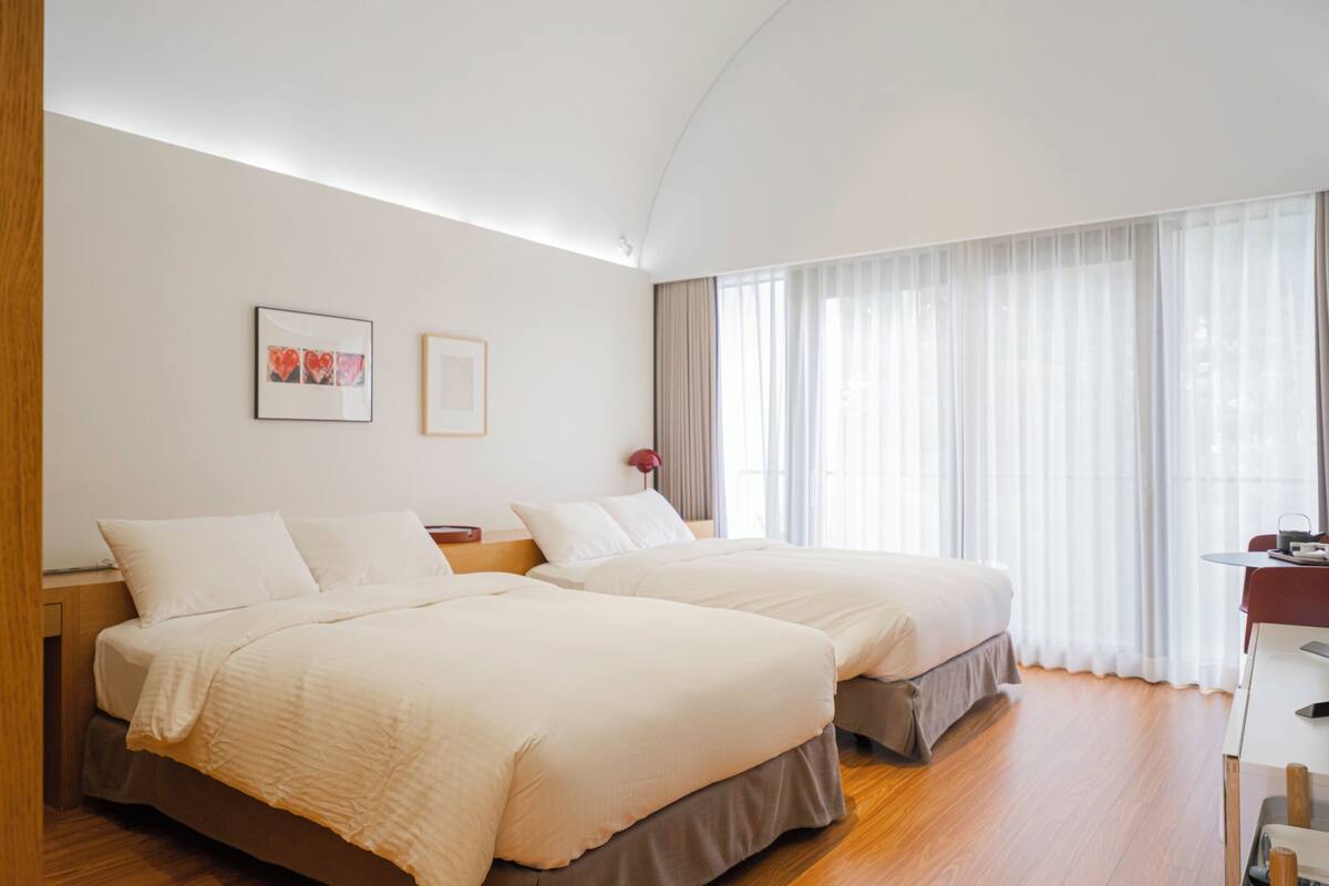 Habitación de hotel con dos camas, un escritorio y un ventanal con cortinas translúcidas.
