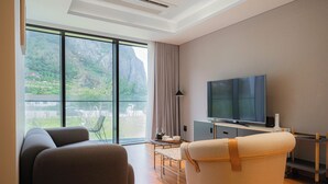 Una sala de estar moderna con un televisor grande, un sofá, una silla y vistas a un paisaje montañoso a través de amplias ventanas.