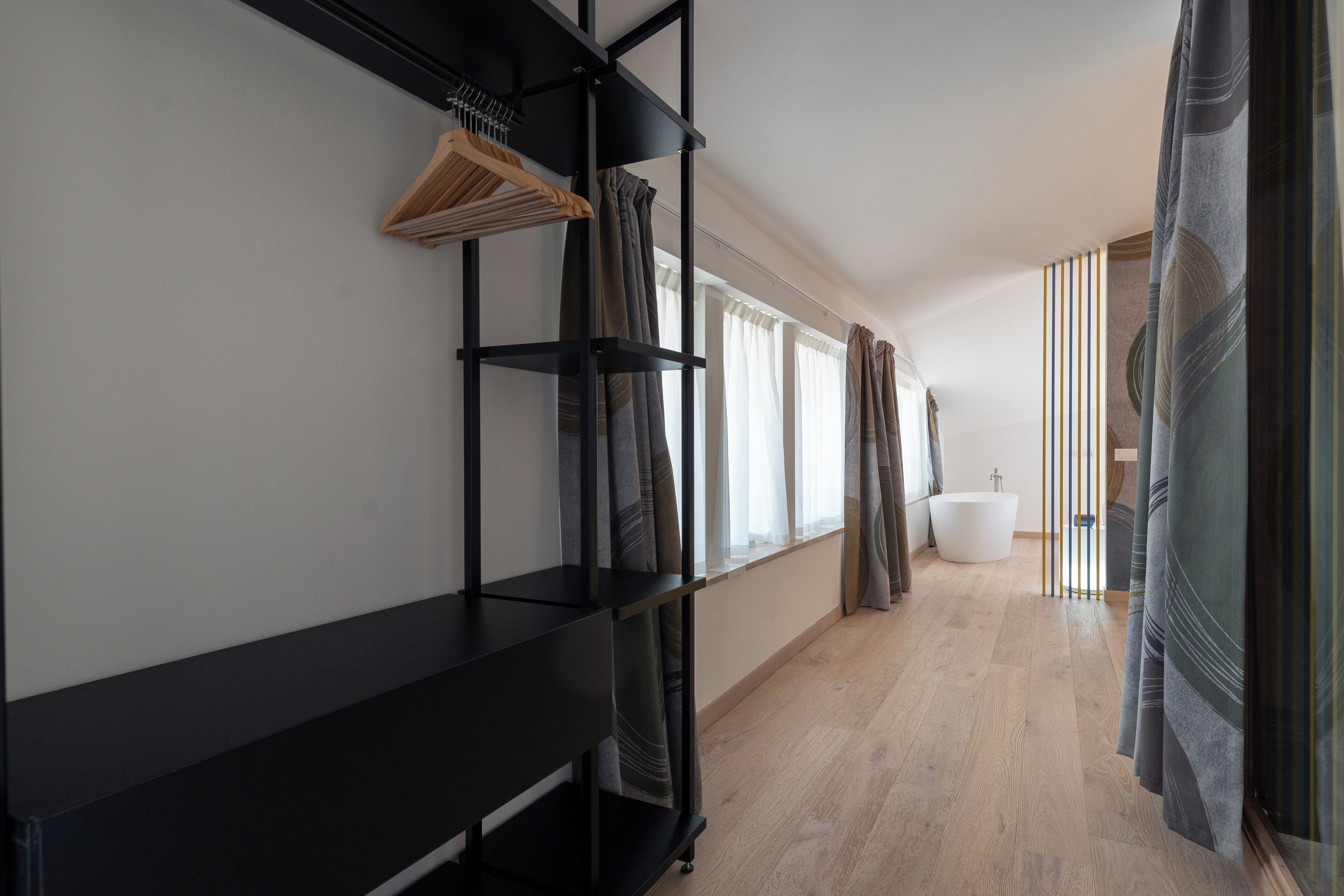 A modern bathroom with a bathtub, wooden flooring, and a black shelving unit with hanging clothes.