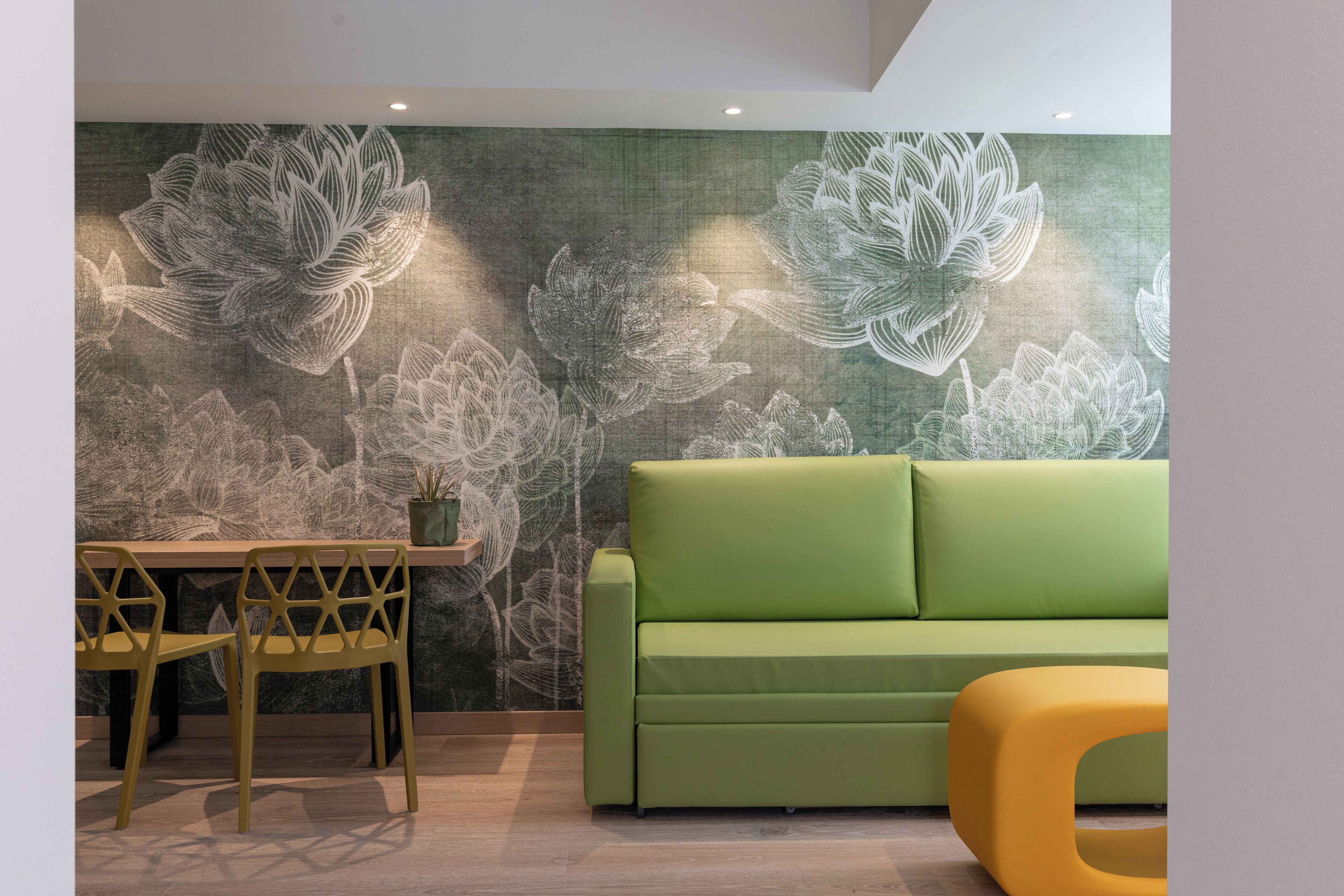 Ein Zimmer mit einem grünen Sofa, einem gelben Hocker und einem Tisch mit Stühlen. Die Wand ist mit blumigem Wandpapier versehen.
