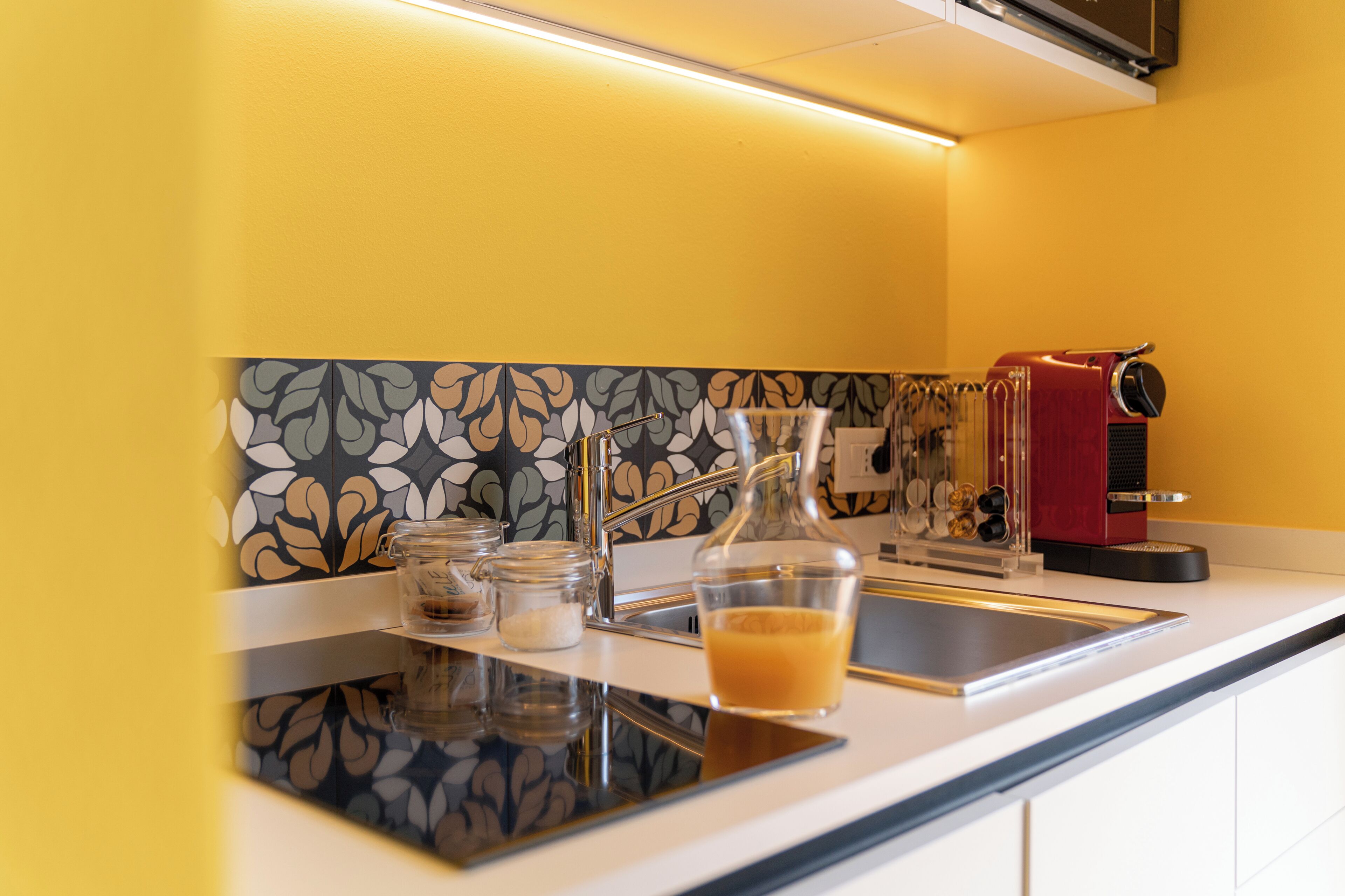 Una cocina moderna con una pared amarilla, un salpicadero con estampado, un lavamanos de acero inoxidable, una cafetera y un vaso de zumo de naranja.