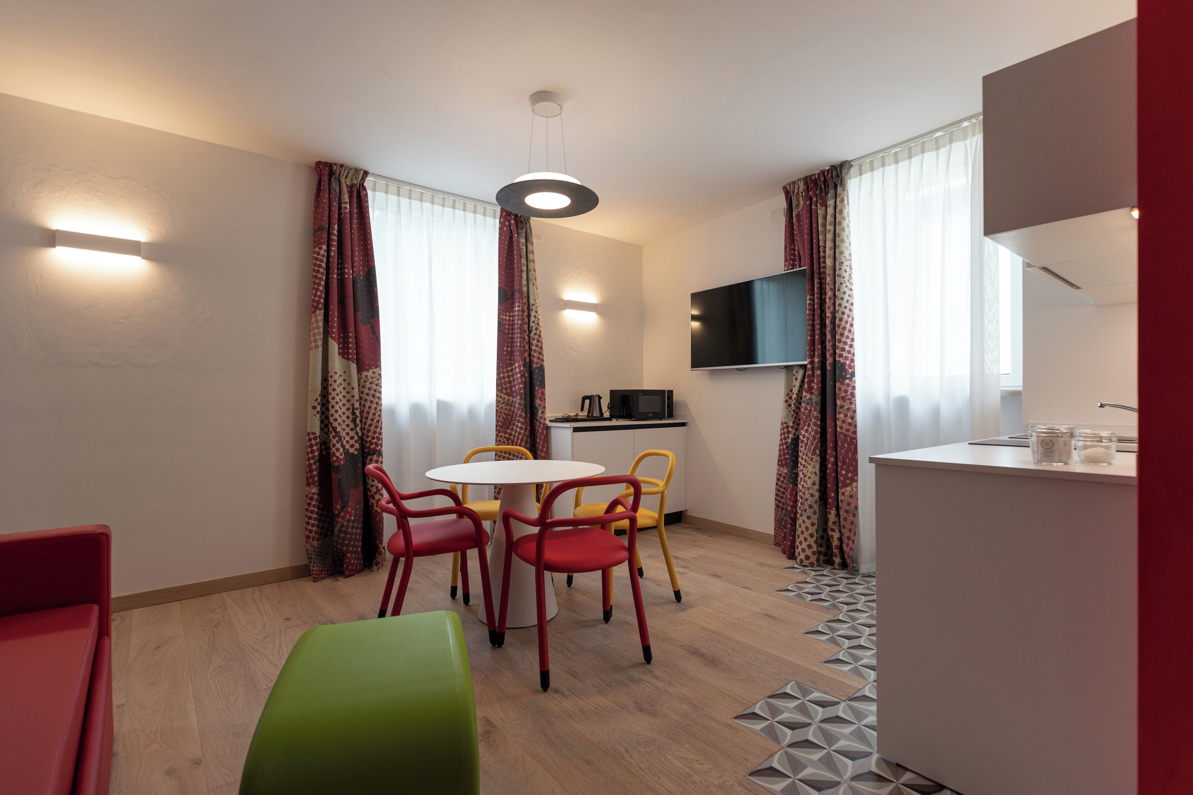 A modern hotel room with a dining area, a kitchen, and a TV mounted on the wall.
