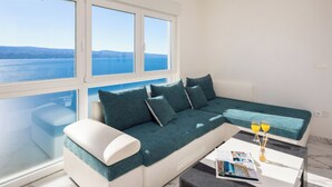 Un soggiorno moderno con un divano bianco e cuscini color petrolio, un tavolino da caffè con riviste e un bicchiere di succo d'arancia, e una grande finestra che si affaccia sul mare e sulle colline in lontananza.