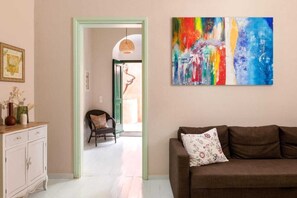 A living room with a brown sofa, a colorful abstract painting, a wicker chair, and a white cabinet. - Traditonal House in Symi (Simi)