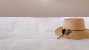 A bed with a white sheet, a straw hat with a flower, and a window with green shutters and a white curtain. - Traditonal House in Symi (Simi)