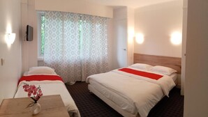 A hotel room with two beds, a small table with a vase, and a window with patterned curtains. - Hôtel les nouveaux Ambassadeurs (Saint-Chamond)