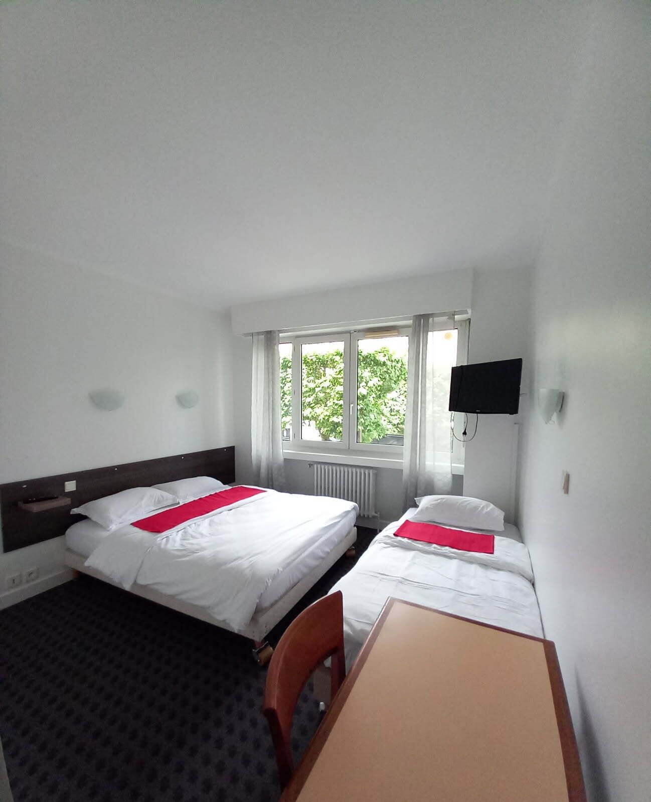 Ein Hotelzimmer mit zwei Betten, einem Schreibtisch, einem Stuhl, einem Fernseher und einem Fenster mit Blick auf Bäume.