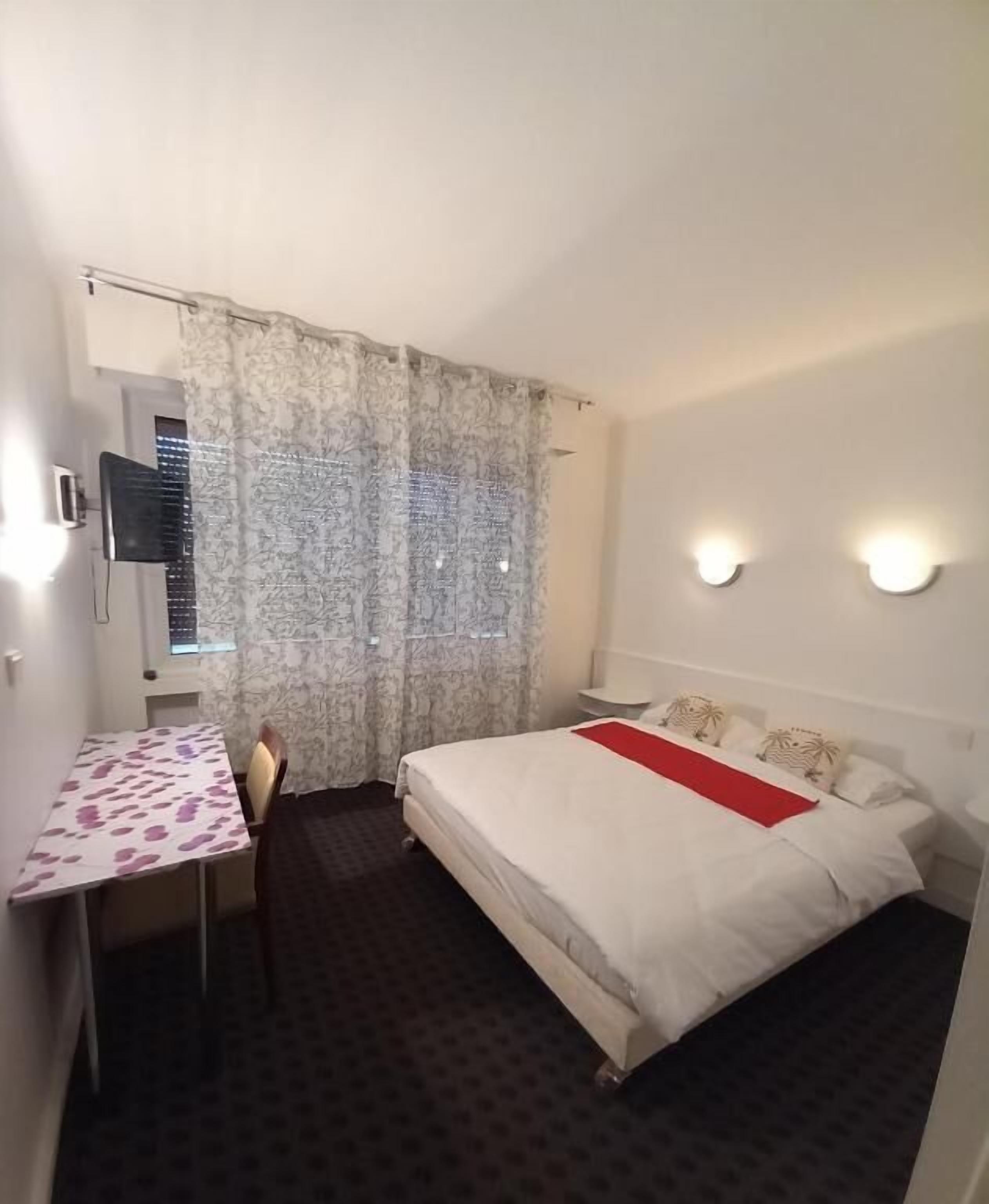 A small hotel room with a bed, a desk, and a chair. There is a window with curtains, a wall-mounted TV, and two wall sconces.