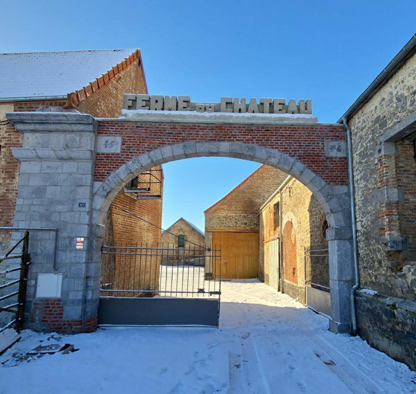 Una entrada arqueada con el letrero "FERME DE CHATEAU" que da a un patio con edificios de ladrillo y una puerta de madera.