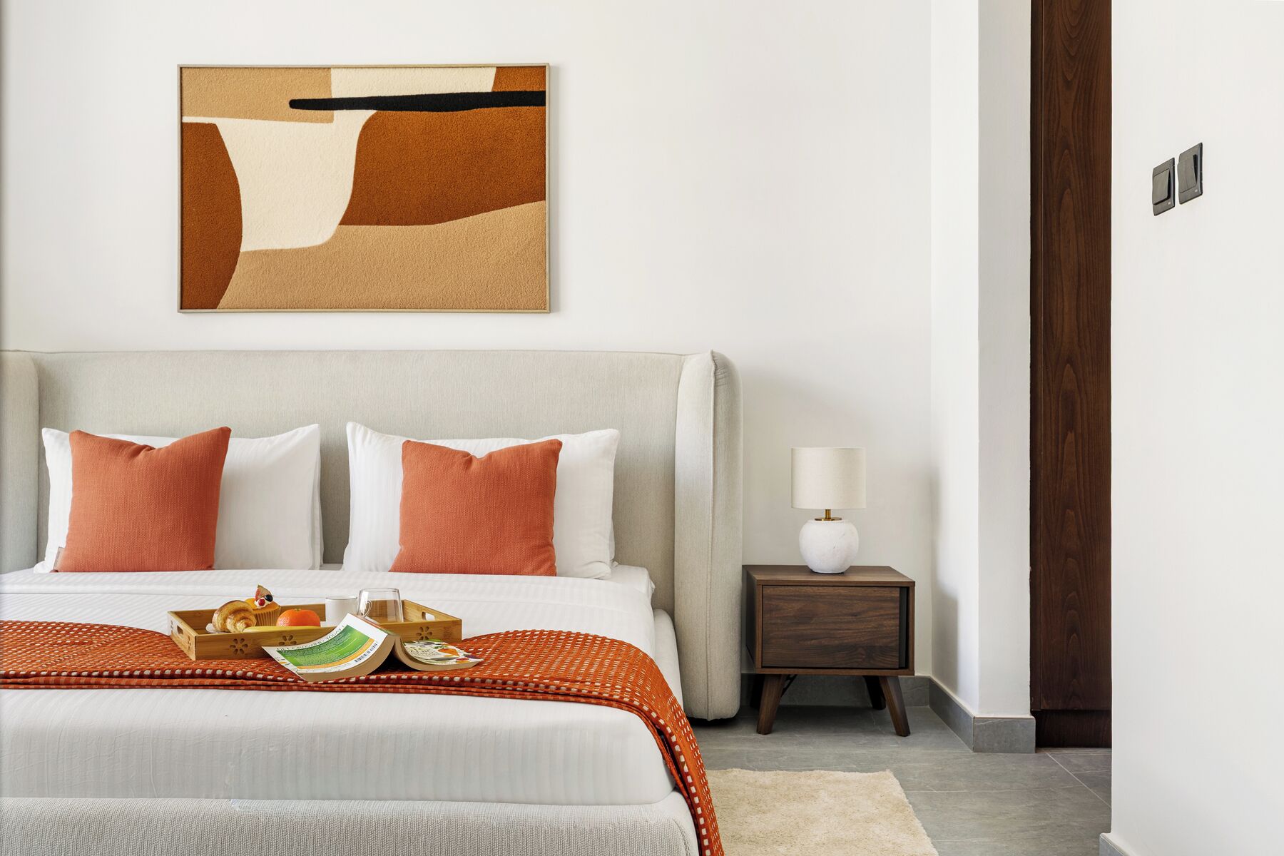 Un dormitorio ordenado con una cama, almohadas, una mesita de noche con lámpara y una obra de arte en la pared.