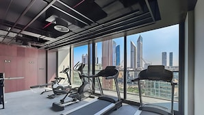 A modern gym with treadmills, stationary bikes, and elliptical machines, featuring large windows with a city skyline view. - 1BR with Huge Balcony Near Burj Al Arab (Dubai)